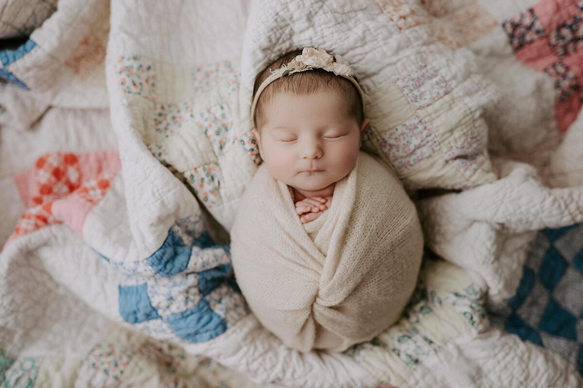 Baby wrapped in a cream knit swaddle with her hands peeking out under her chin. Baby is laying on an heirloom vintage quilt.
