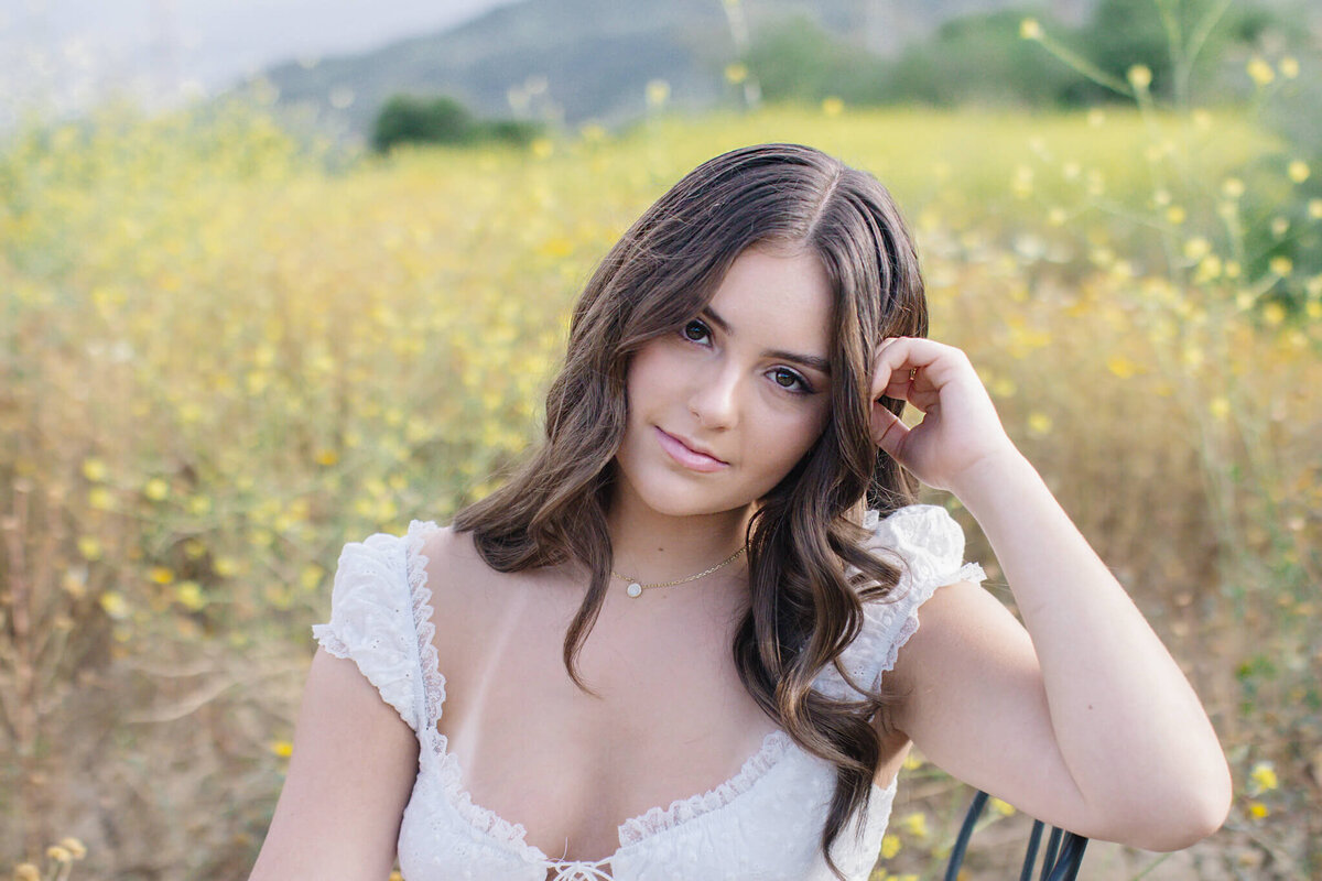 A woman with long, wavy brown hair sits in a field of yellow flowers. She is wearing a white, lace-trimmed dress and has a gentle expression, with one hand resting on her head. The background shows blurred greenery and distant hills.