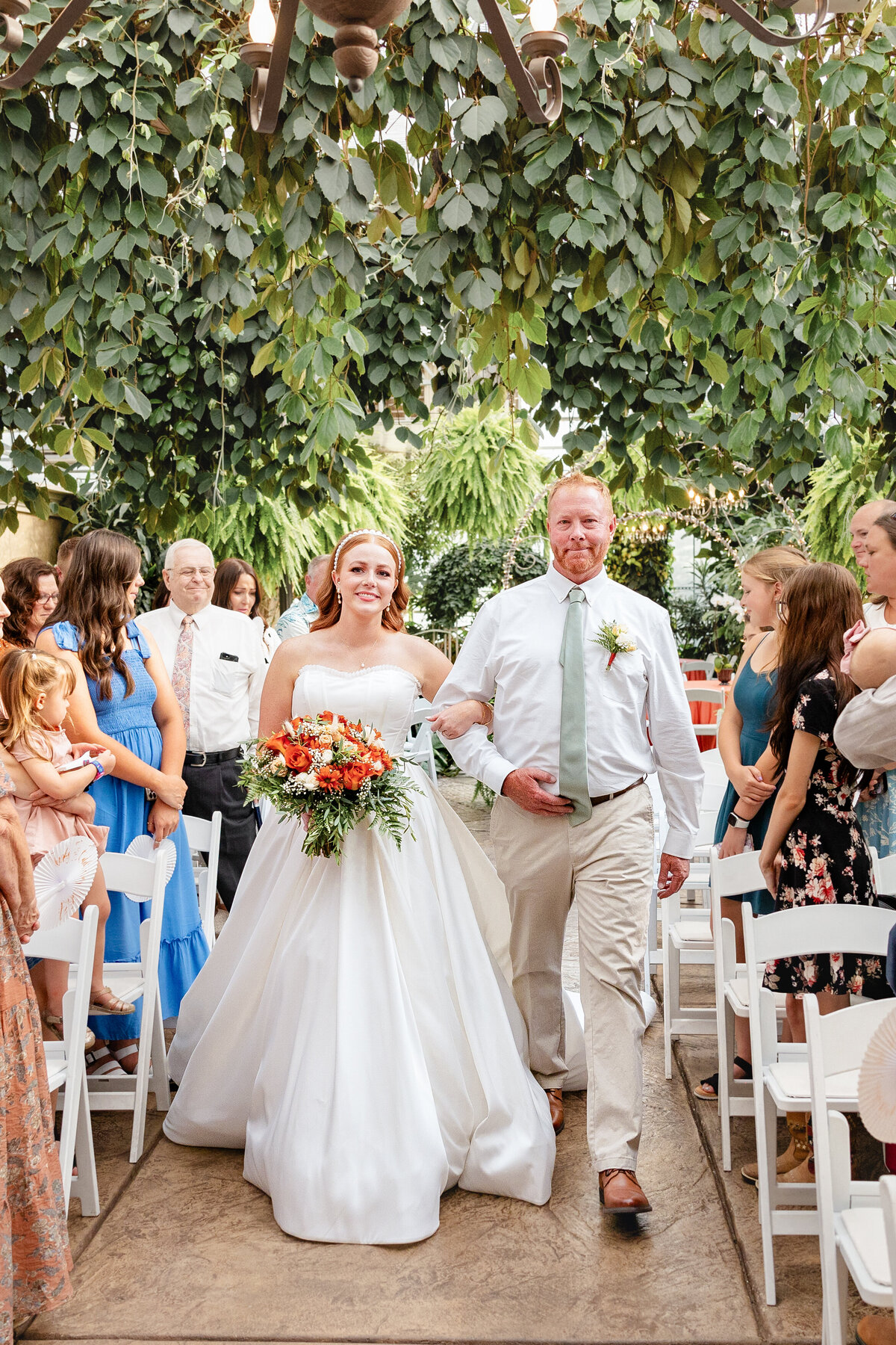 The bride, holding a bouquet of orange and white flowers, walks down the aisle with her father. Guests on either side smile as they watch her approach the groom.