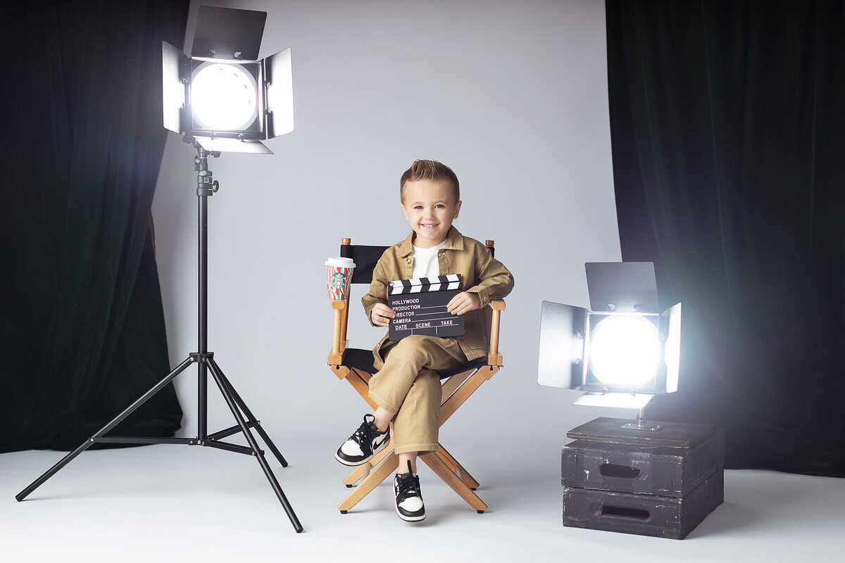 Young boy in a tan outfit sits in a director’s chair holding a Hollywood clapboard, surrounded by studio lights in a cinematic Dallas milestone photographer setup.