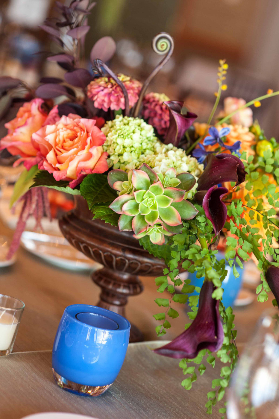 fall centerpiece of orange roses, succulents, purple calla lilies, ferns, green amaranthus, zinnia, fiddlehead fern in brown urn with blue glassybaby candle