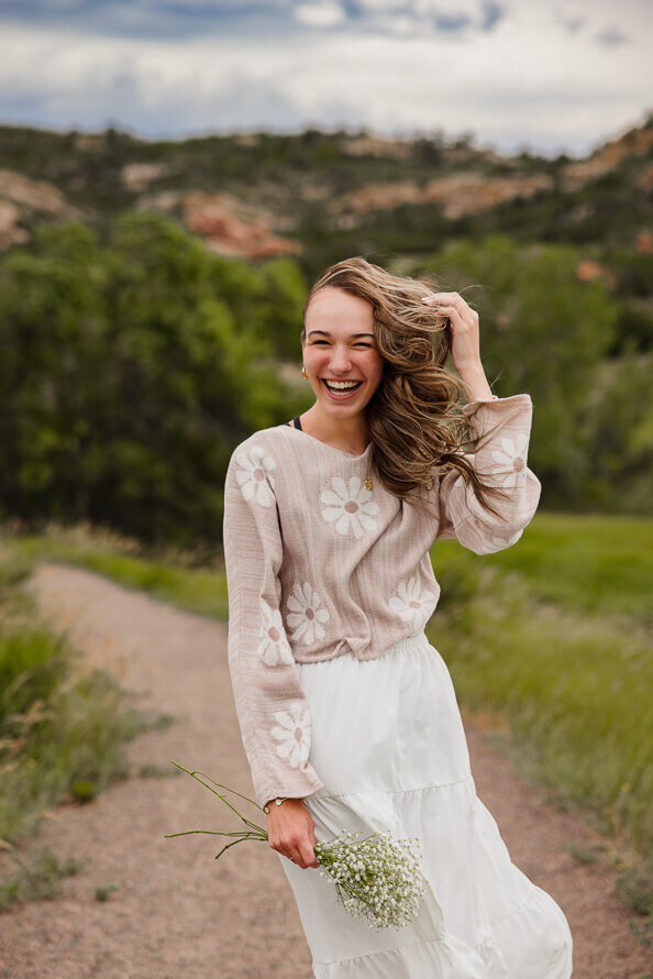 Girl in white skirt and tan sweater laughs as wind blows her hair during a photo session with Angela Marie Media.