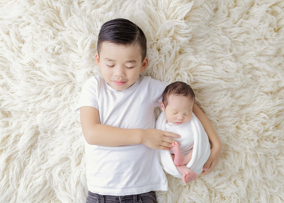Older brother holding newborn sibling on creamy fur rug - neutral studio session Maple Grove