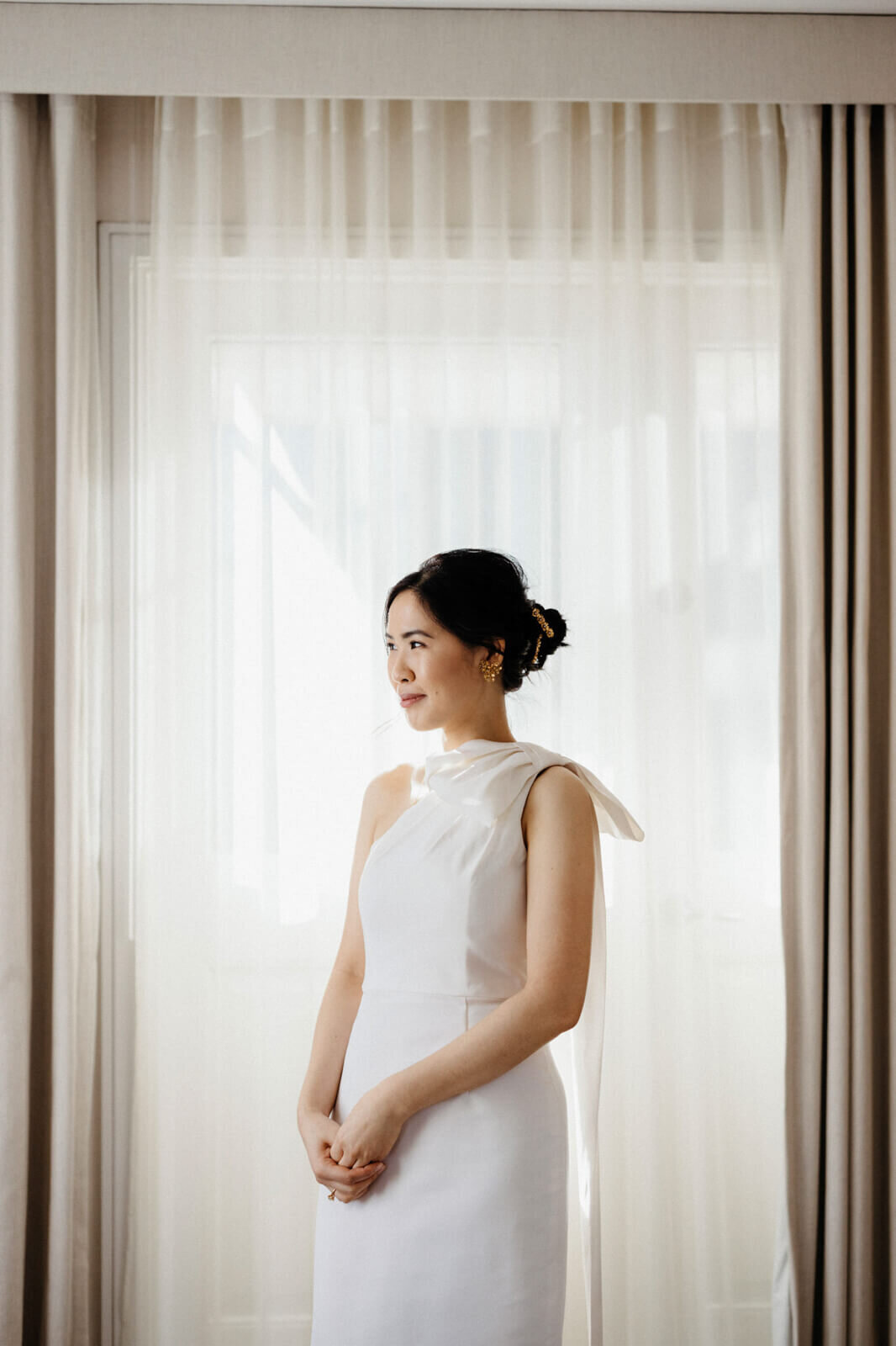 The bride, whose hands are clenched together, is standing while looking sideways in front of a  window with an off-white curtain