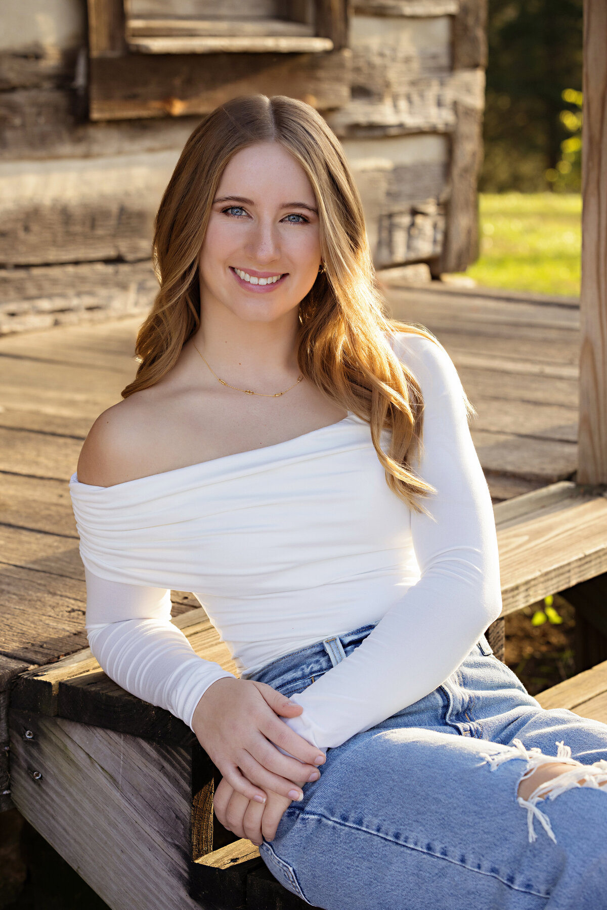 Smiling high school senior in jeans and white top seated on wooden steps of a rustic cabin at Anne Springs Close Greenway