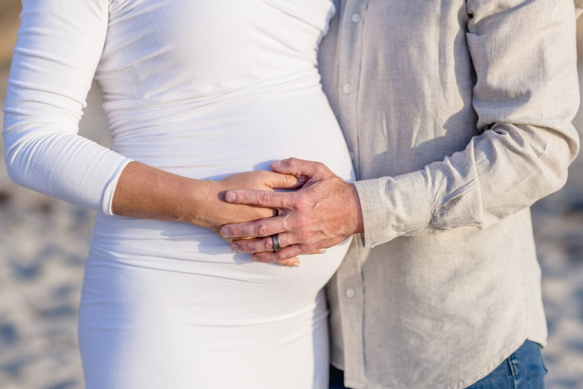A couple embracing, with the persons hands gently resting on a pregnant belly. One is wearing a white dress and the other a light-colored shirt. The background is a sandy area, suggesting a beach setting.
