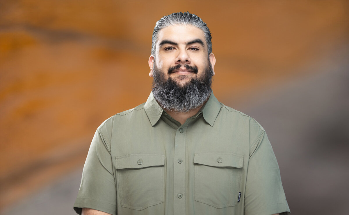 Professional headshot of a bearded man from Messer Construction, wearing a sage green button-up shirt, with a calm and confident expression, photographed at Janel Lee Photography studio in downtown Cincinnati, Ohio. He stands against a warm, softly blurred orange and gray background that creates a modern yet approachable feel.