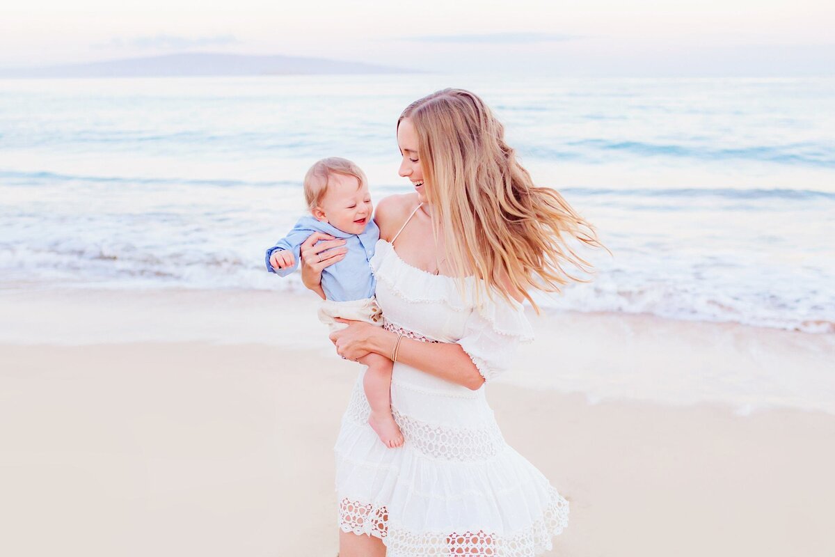 Mom with long blonde hair and white dress twirls her young son on her hip while being photographed by Love + Water at sunrise on Maui