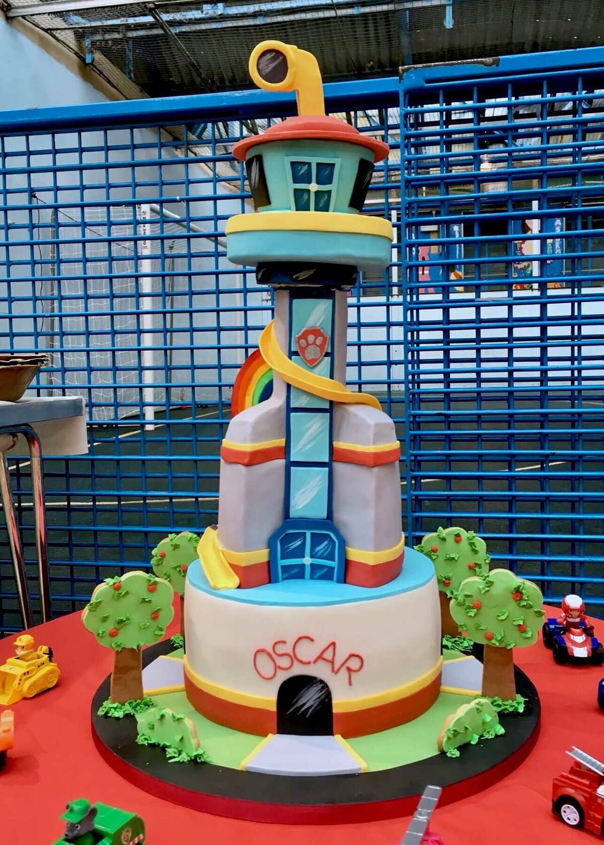 An elaborate very tall cake of a tall building with a slide running around the building and a periscope coming out of the top with a Paw Patrol theme