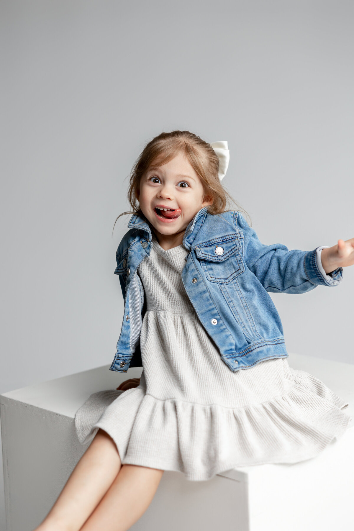 Little girl laughing in light blue denim jacket and cream dress, sitting on cube in studio. Photographed by Yucaipa family photographer Kaitlyn Dawn Photography.