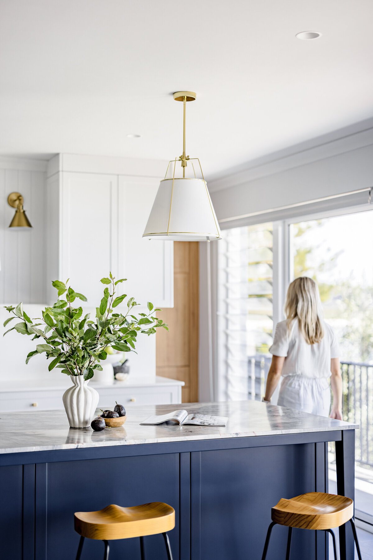 Modern kitchen with a marble island, two wooden stools, a vase with greenery, and a person contemplating the view—exemplifying bespoke interior design craftsmanship in Newcastle NSW.