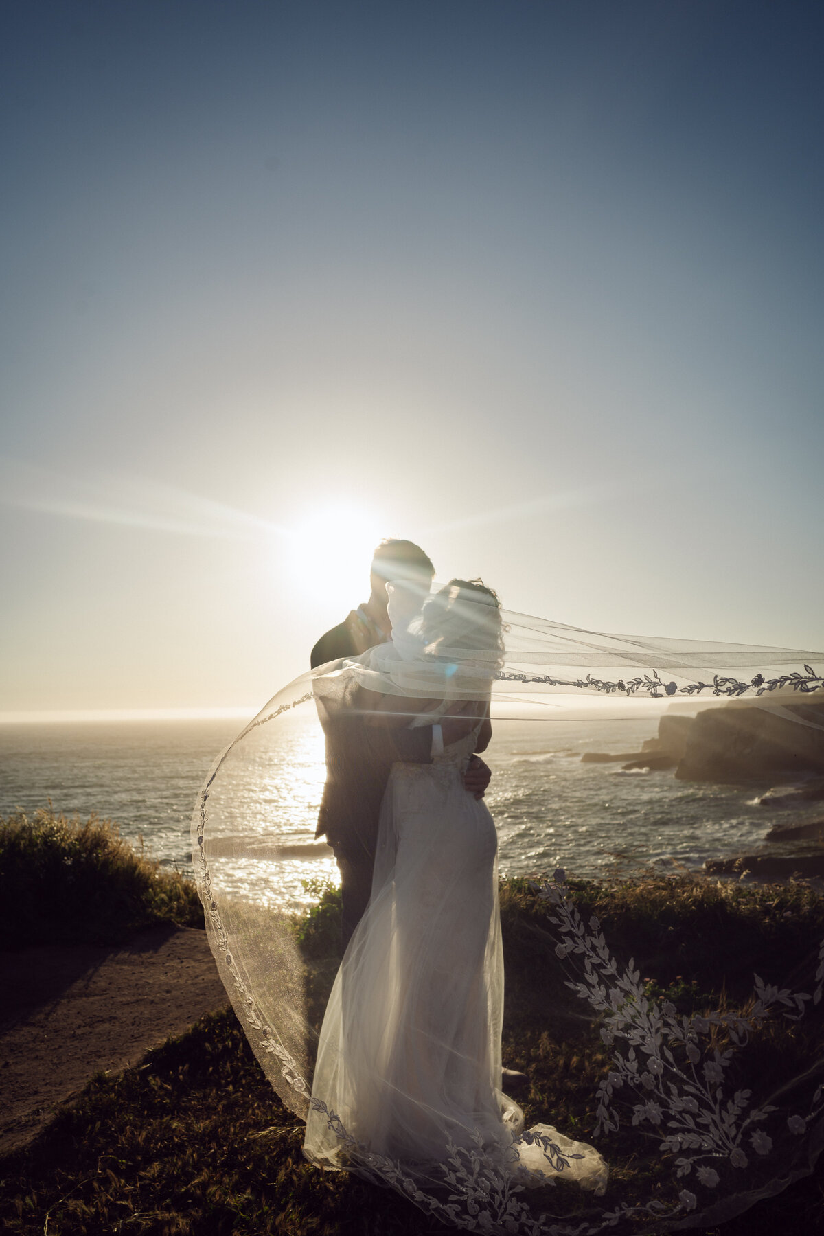 Bride and groom elopement at Shark Fin Cove cliffs Santa Cruz during golden hour with her Claire Pettibone veil blowing in the breeze 