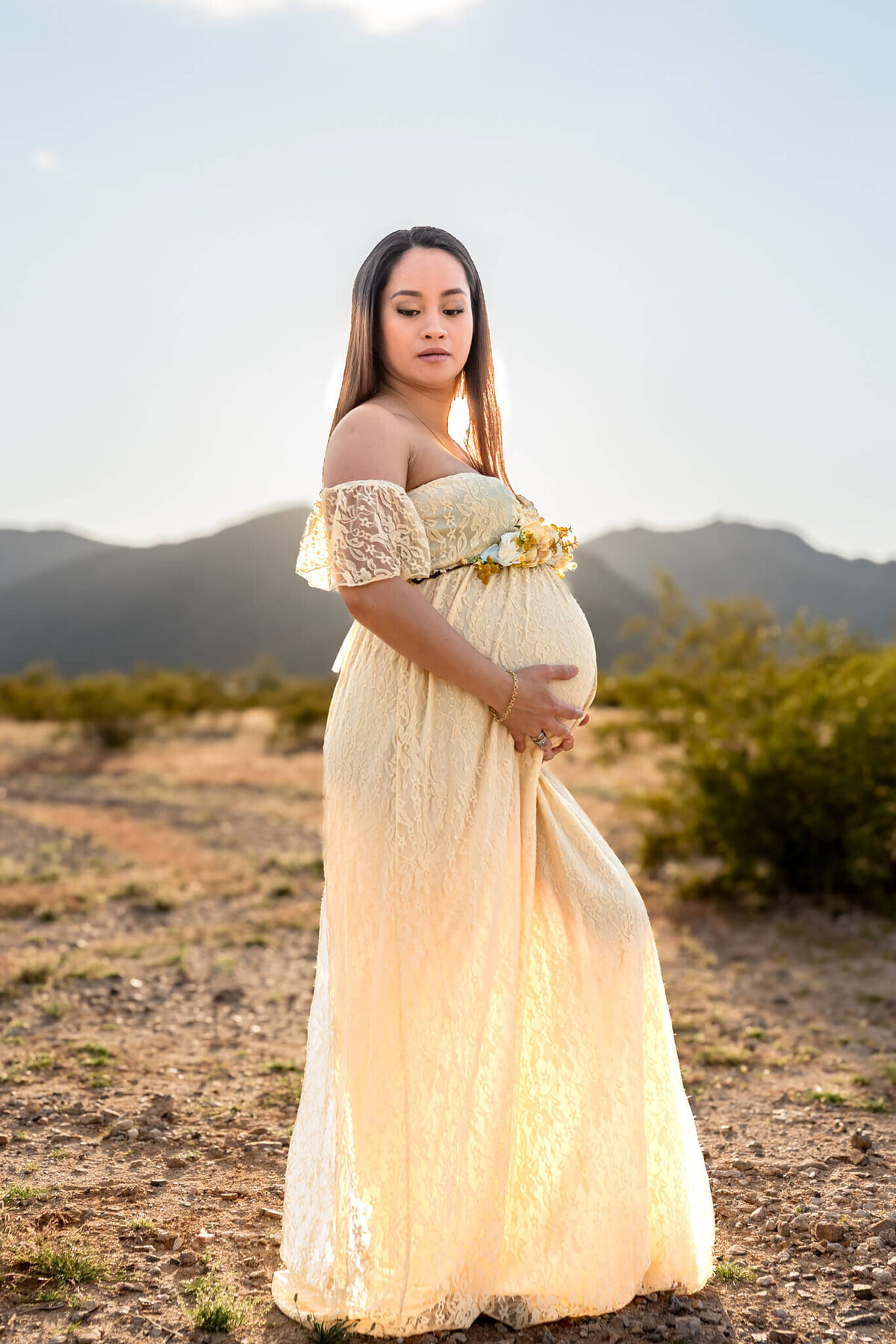 Mother-to-be in cream color lace dress holding her baby bump with the sun behind her