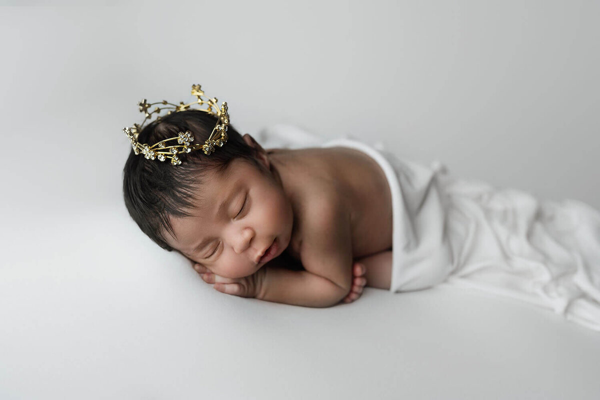 A newborn baby in a golden crown and covered by a white blanket sleeps on a studio pad posed by a New Orleans Newborn Photographer