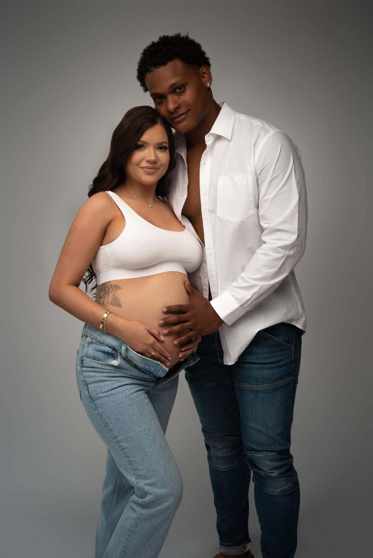 Man in white button down shirt and jeans posing with pregnant brunette woman wearing white sports bra and light jeans on grey backdrop