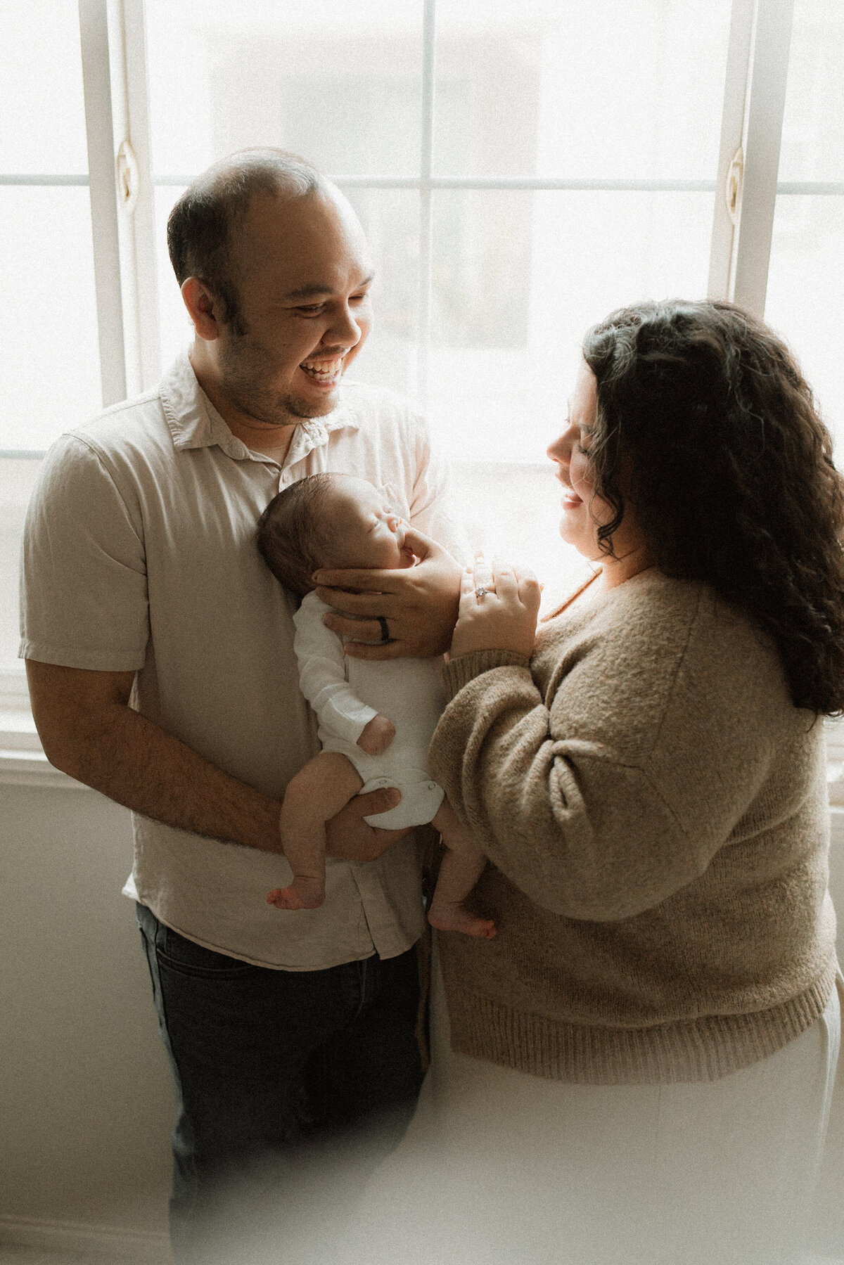 Father cradling his newborn in soft light, smiling at mom, who's standing right next to him.