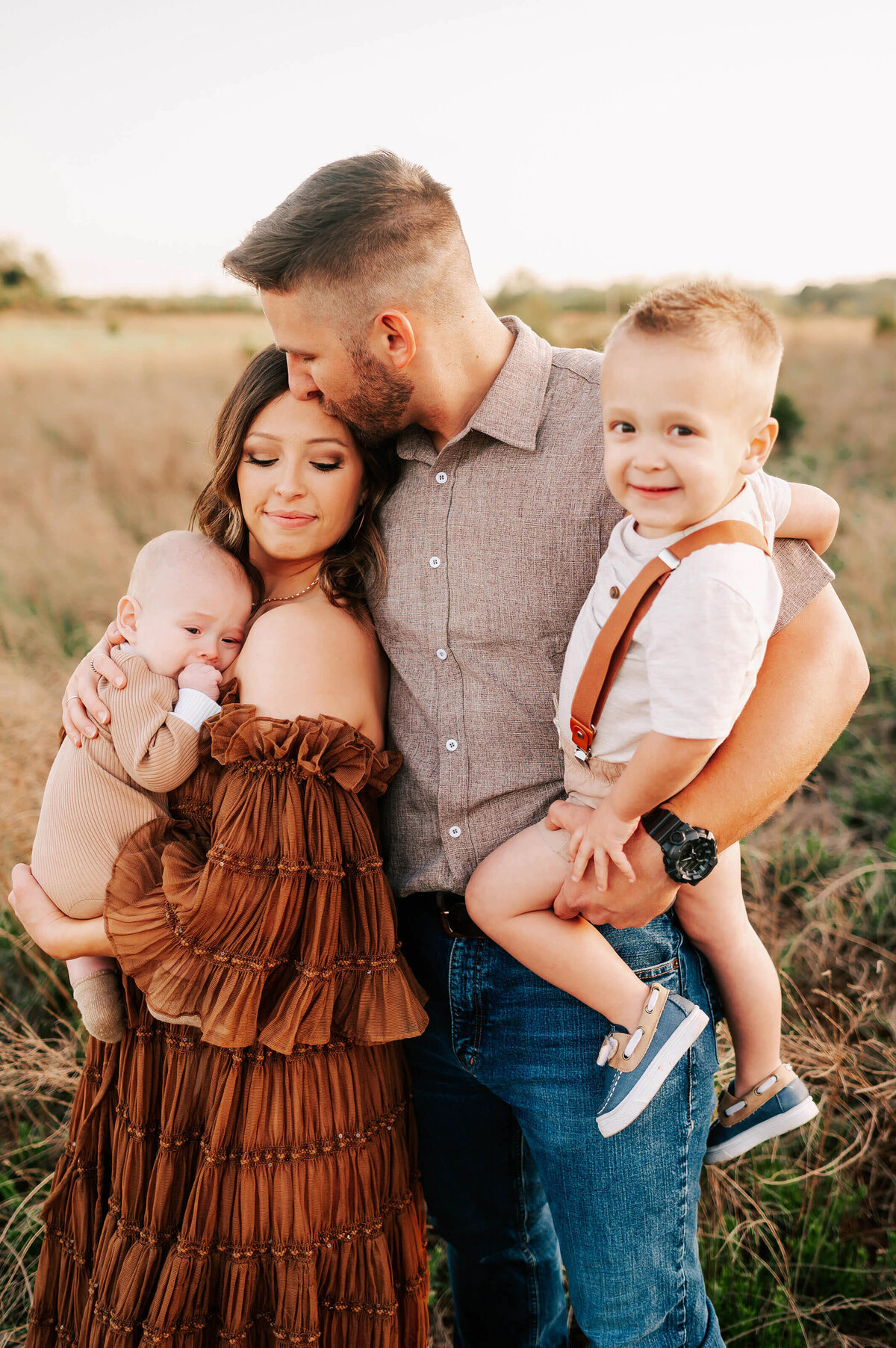 husband kissing wife holding kids in a field  during Springfield MO family photography session