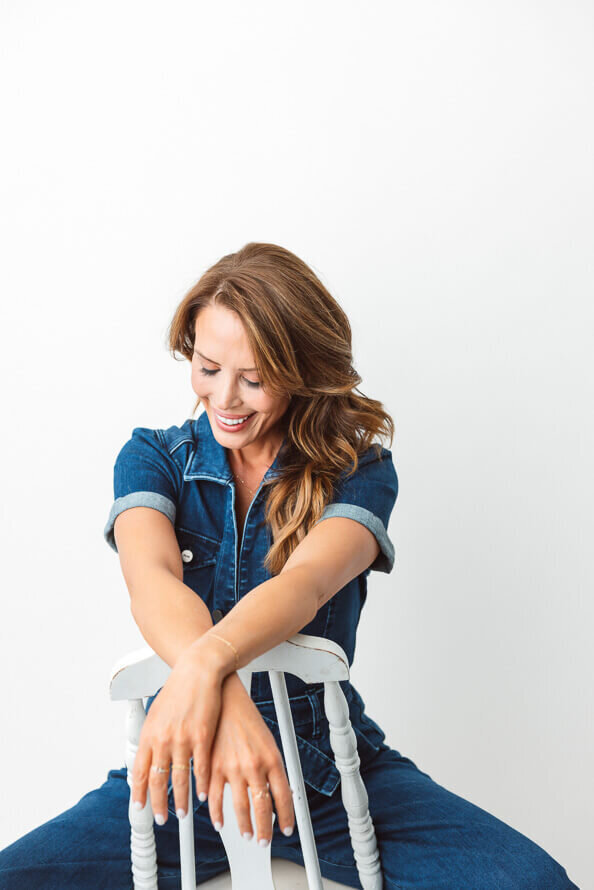 Girl in jean shirt sits backwards on a white chair and looks down smiling.