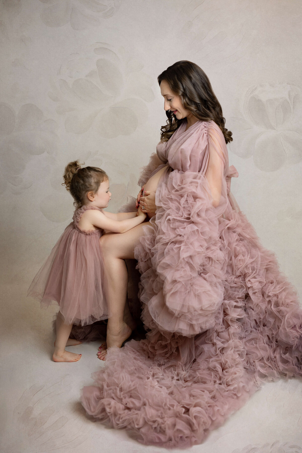 pregnant mother in a fluggy pink maternity gown sitting while her daughter is wearing a matching dress and holding her pregnant belly in a photography studio
