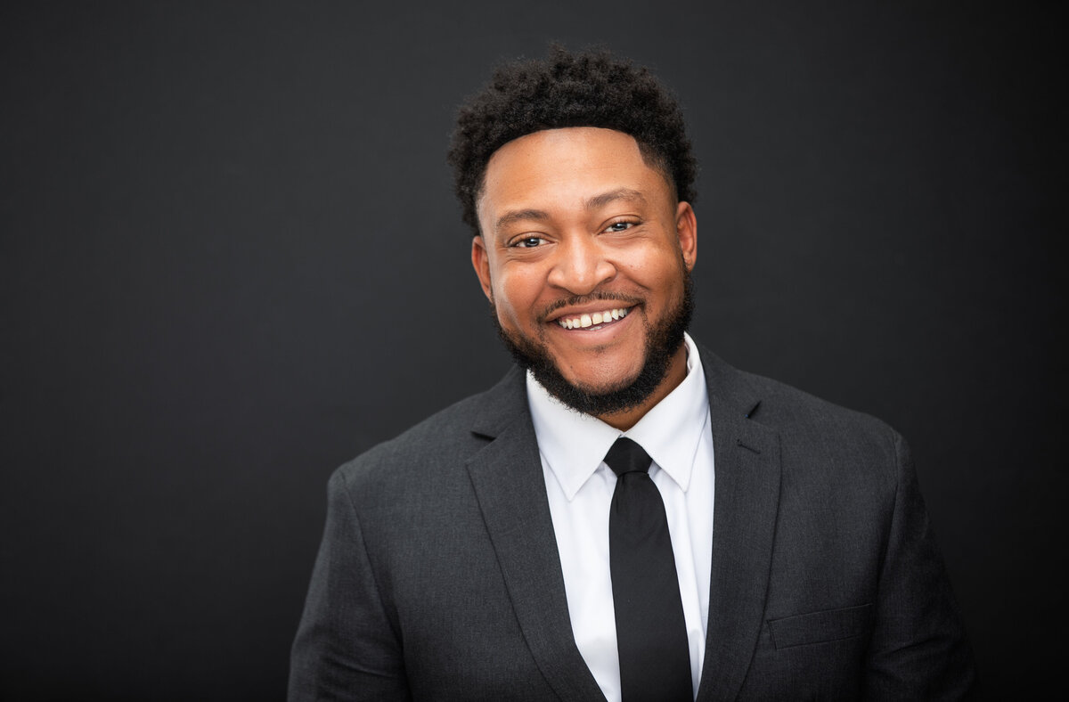 An African American black businessman in a black suit and black tie smiles and poses for a professional headshot photo at Janel Lee Photography studios in Cincinnati Ohio