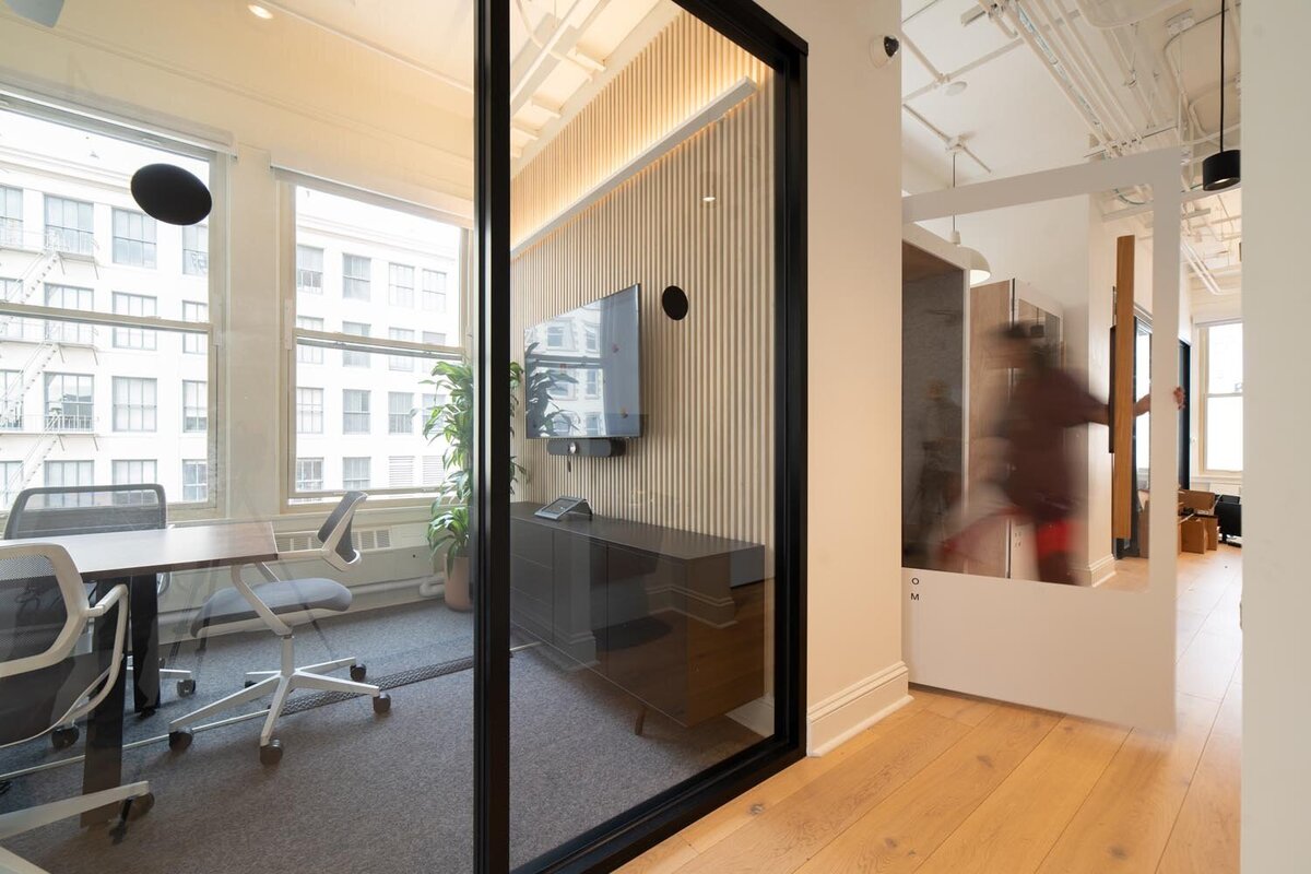 Modern glass-enclosed meeting room with wood slat accent wall, mounted TV, and white office chairs, with someone entering a phone booth in the background.