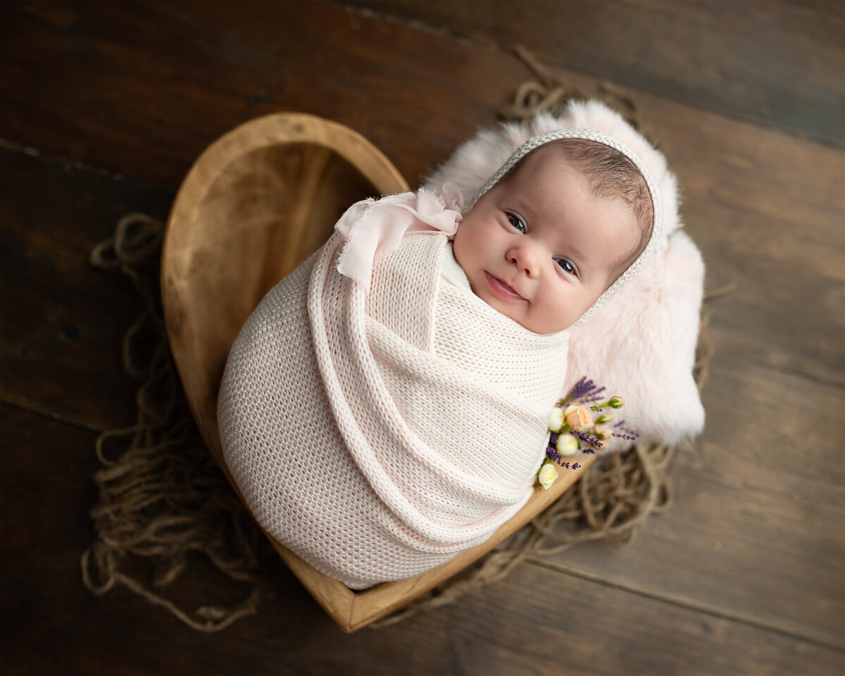 Heartfelt client testimonial for newborn photography featuring a swaddled baby in a pink wrap, nestled in a handcrafted wooden heart bowl with floral details. Captured in a warm, rustic-styled photography session at Meraki Photographic Creations Studio in Paris, Ontario.