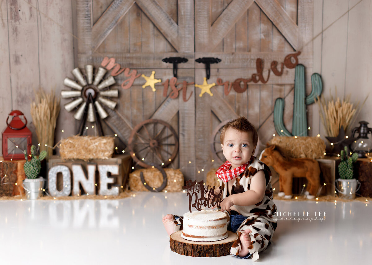 Rustic My First Rodeo, Rodeo themed cake smash in West Palm Beach, FL newborn and cake smash photography studio.  Baby boy wearing cow print and is sitting with white cake between his legs with a fist full of cake looking at the camera. In the background, there are hay stacks, cactus plants, horseshoes, lanterns, plush horse and wagon wheels.
