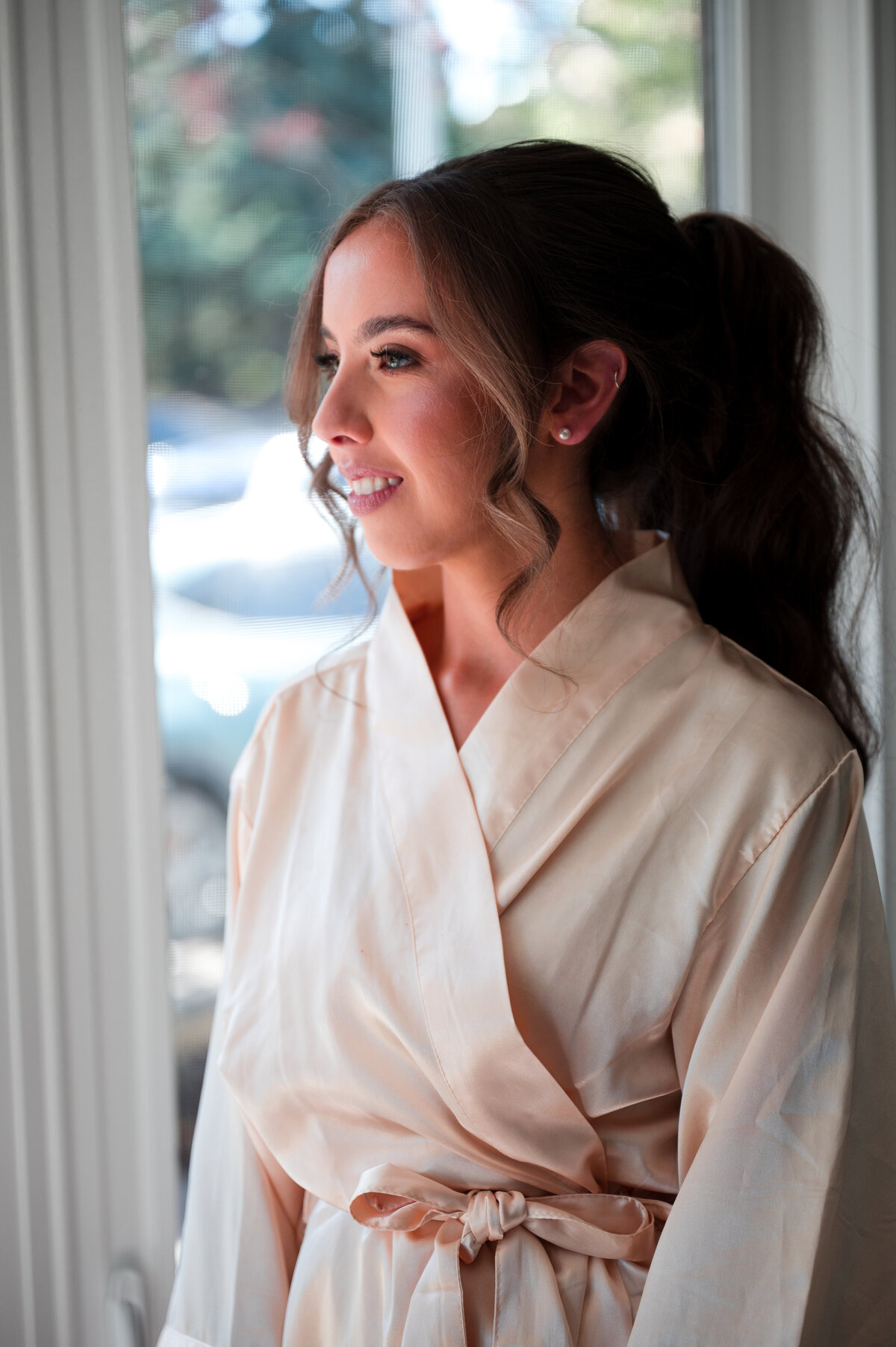 bride standing near windows in a white robe, finishing getting ready for her wedding day