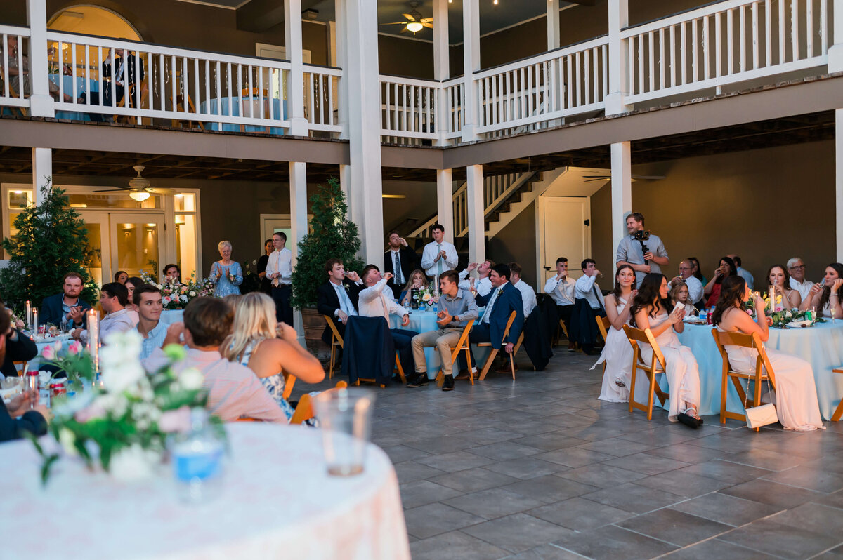 A group of wedding guests in formal attire sit at round tables outdoors under a large, two-story veranda, attentively watching a speaker during a reception.