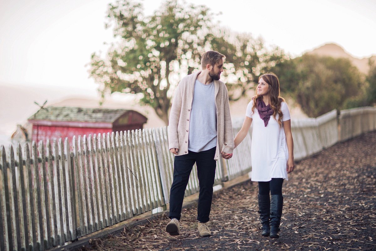 Engaged couple go for walk holding hands and talking along a white picket fence during an engagement session
