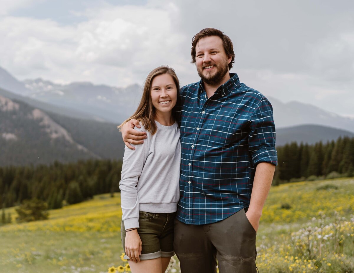 Alpine Vows, a Colorado elopement photographer and videographer team, standing together in a mountain meadow with wildflowers, capturing authentic and adventurous wedding stories.