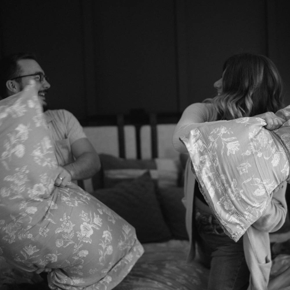 A couple laughs during a pillow fight during their playful maternity session in Spokane, Washington, highlighting joy and spontaneity.