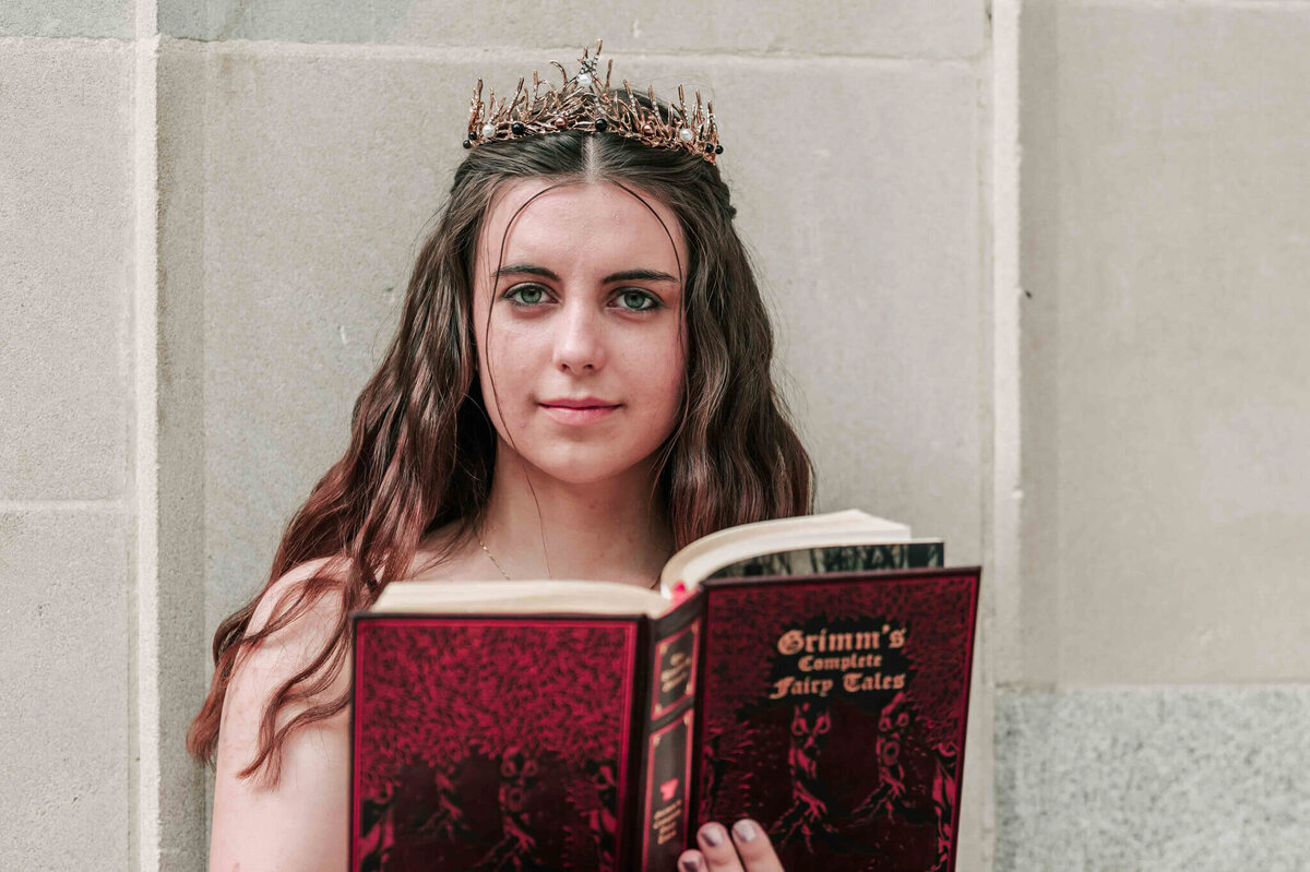 A brunette girl wearing a tiara and holding a book.