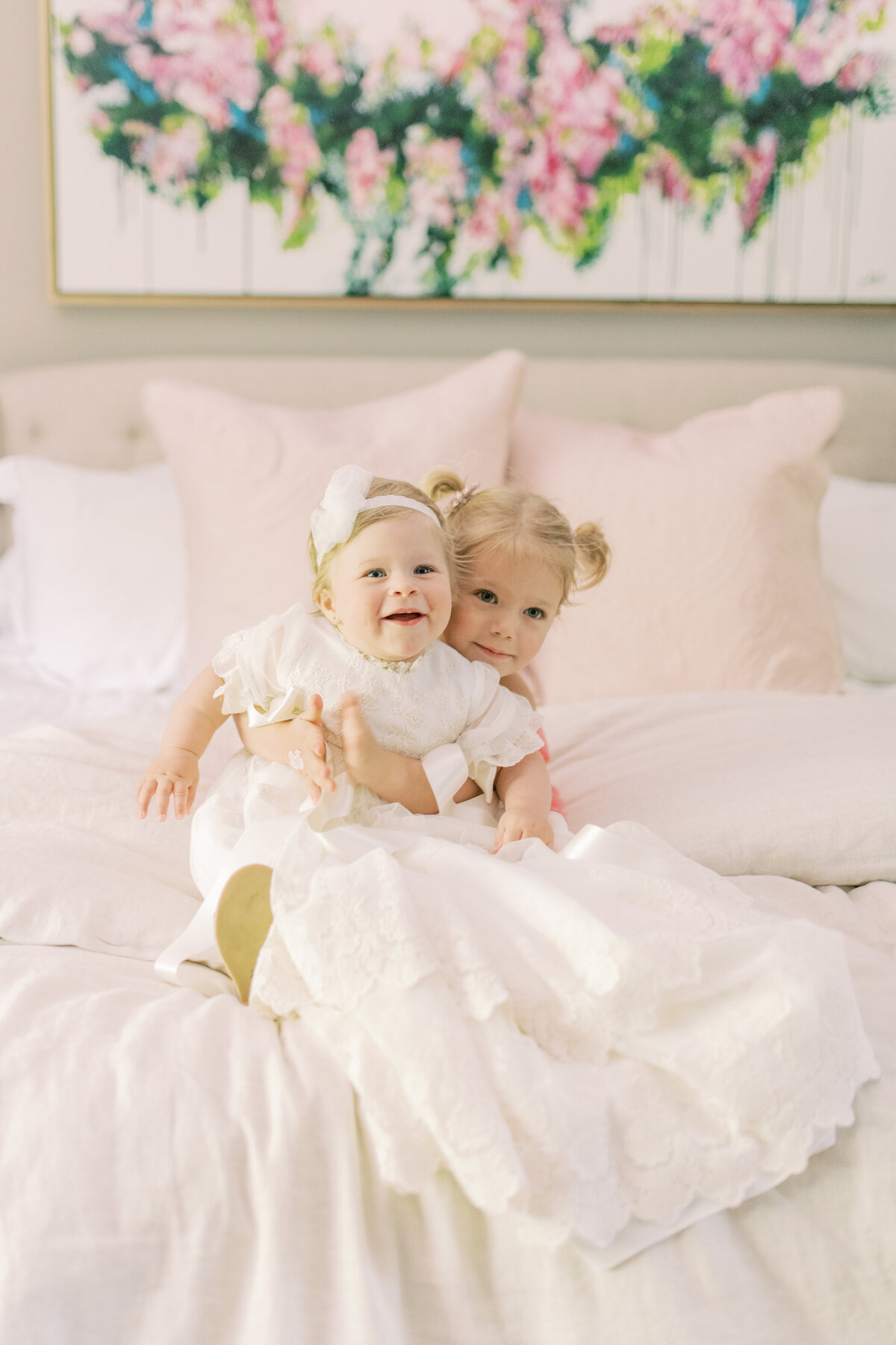 Two sisters embrace on crisp white bedding, one in a flowing christening gown, echoing legacy and love.