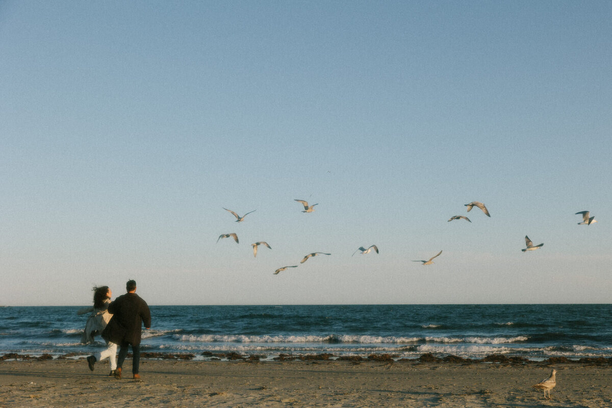 Couple running on the Newport Beach with seagulls flying around them in Rhode Island, captured by Andrea-Marie Photography