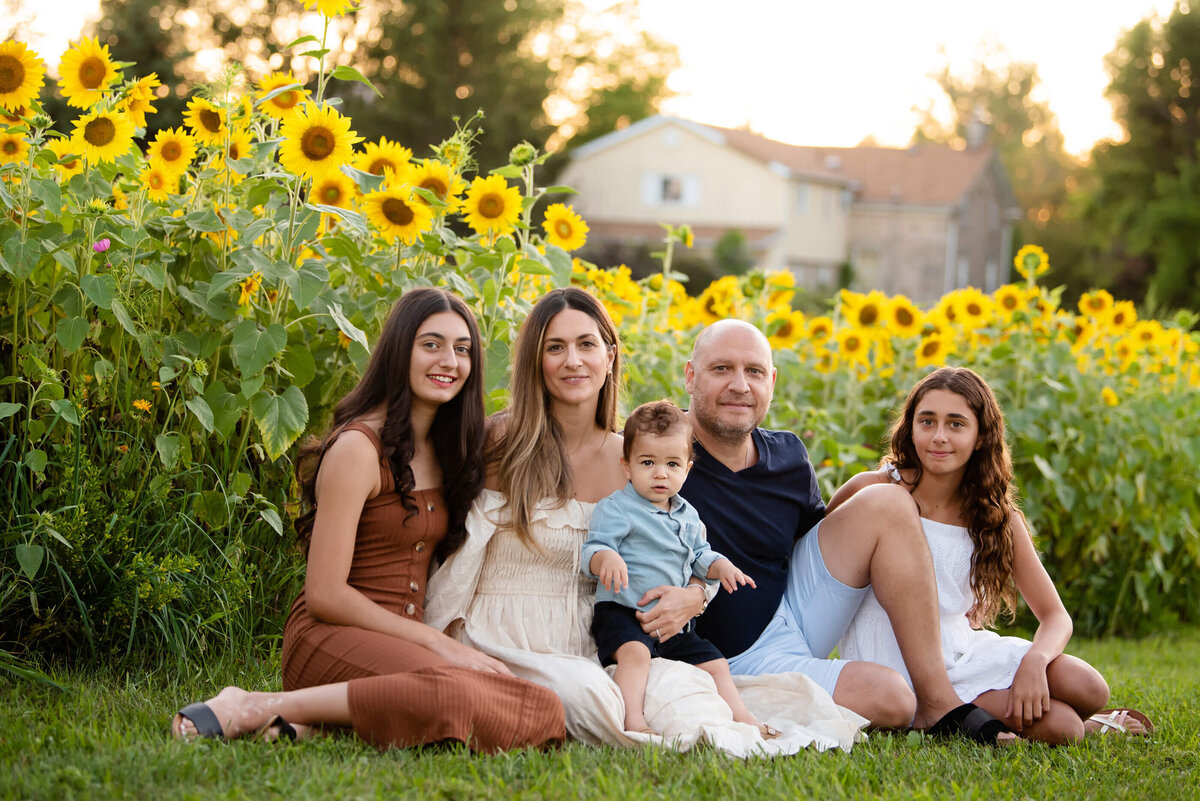 mom and dad sitting on the ground with their two teenage daughters and toddler son in front of a field of sunflowers for their spring Burlington family photos