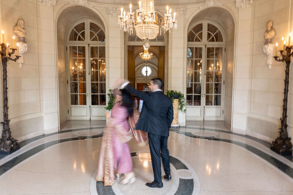 A couple is dancing in an elegant hall with marble floors and chandeliers. The man wears a dark suit and the woman is in a pink traditional dress. Motion blur adds a sense of movement to the scene. Arched windows and decorative statues adorn the background.