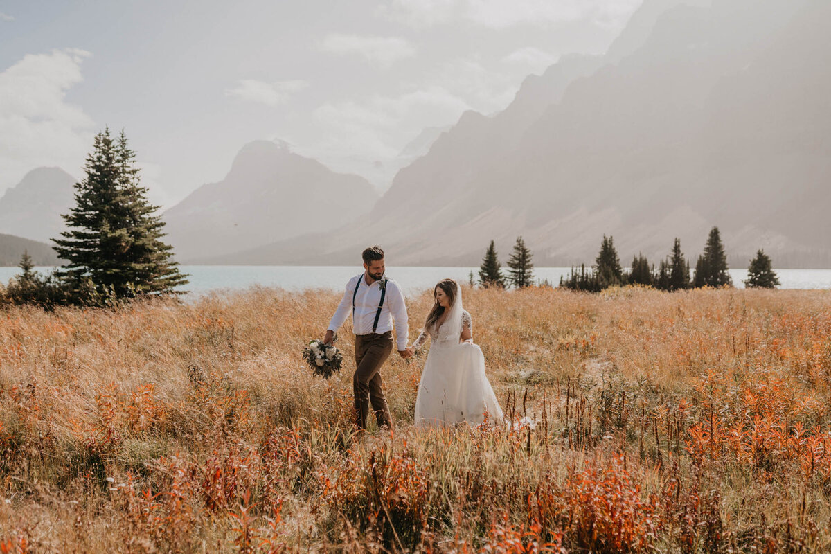 Bride and groom walking hand in hand through a gorgeous field in the mountains of Alberta, fall wedding inspiration, captured by Ash Maclean Photography, romantic elopement and wedding photographer in Red Deer, Alberta. Featured on the Bronte Bride Vendor Guide.