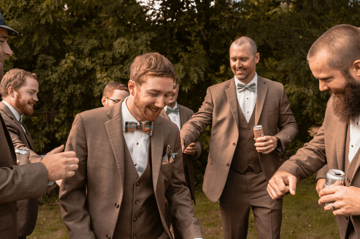 A candid photo of a groom and his groomsmen laughing and celebrating together outdoors. The group is dressed in brown suits with bow ties, enjoying a lighthearted moment before the wedding ceremony.