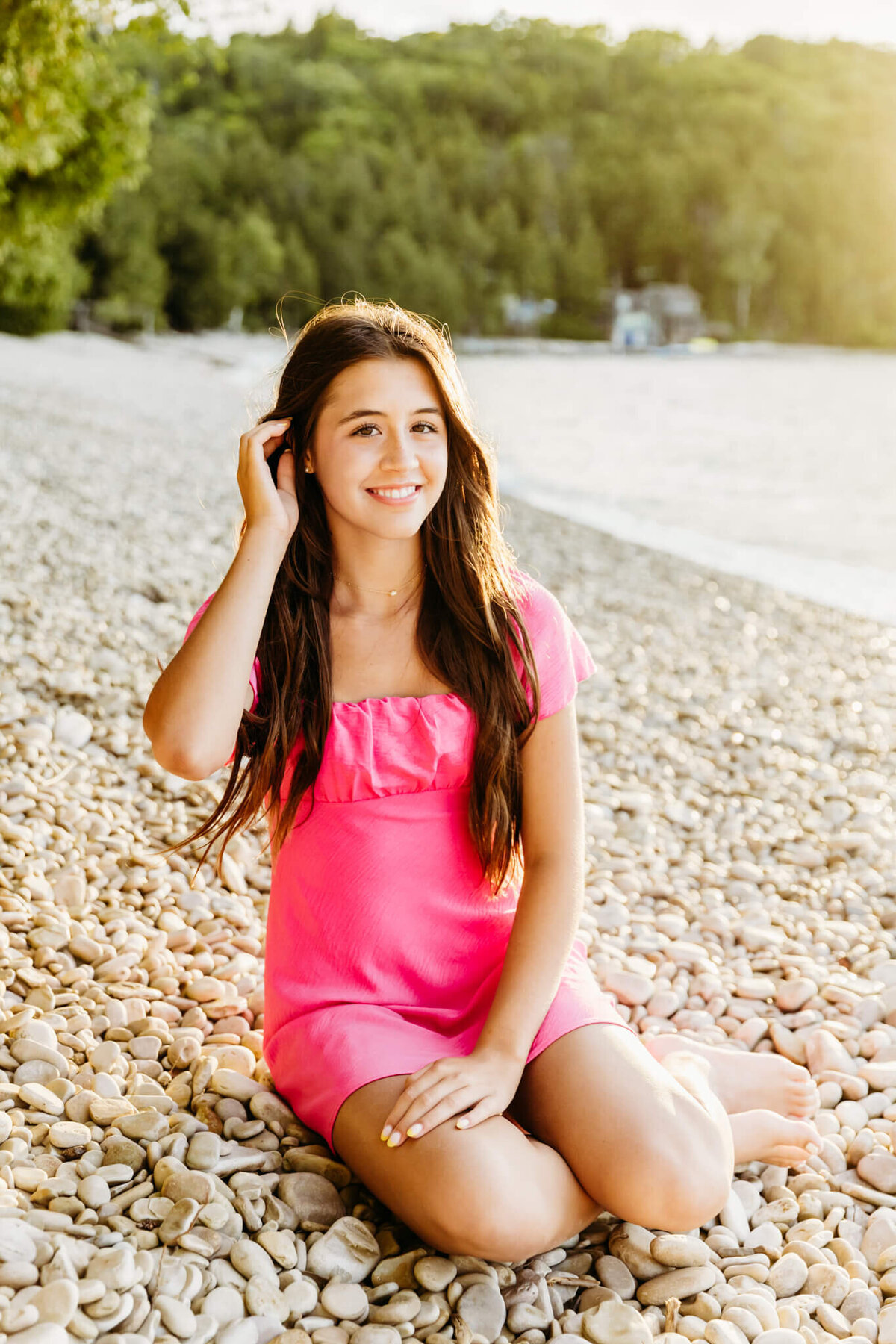 pretty senior girl in a short pink dress sitting on a rocky beach tucking hair behind her ear near Oshkosh