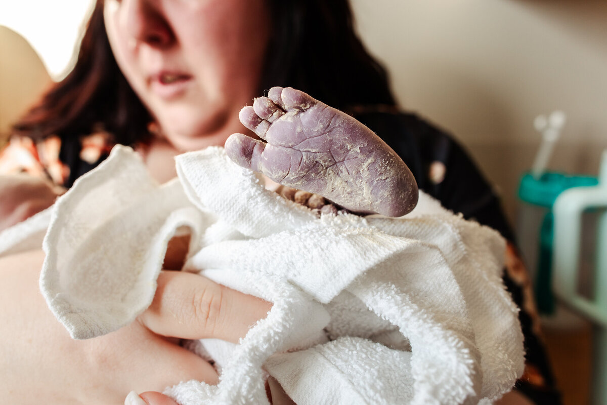 A mother holds her newborn baby, and his foot sticks out of the towel, covered in white vernix