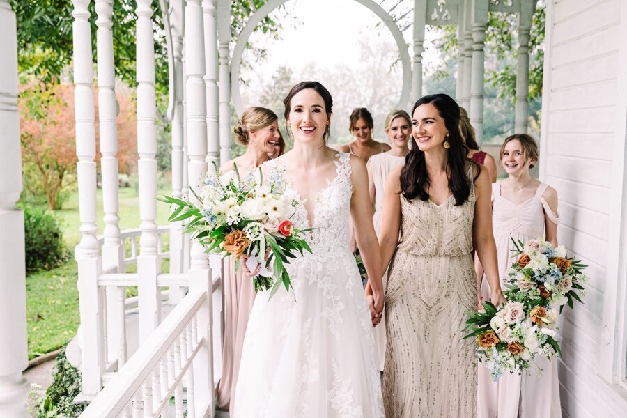 A joyful bride holding a romantic white and blush bouquet walks hand-in-hand with her bridesmaid, surrounded by her wedding party on a charming, wraparound porch. Lush greenery and soft pastels set a fresh, springtime tone