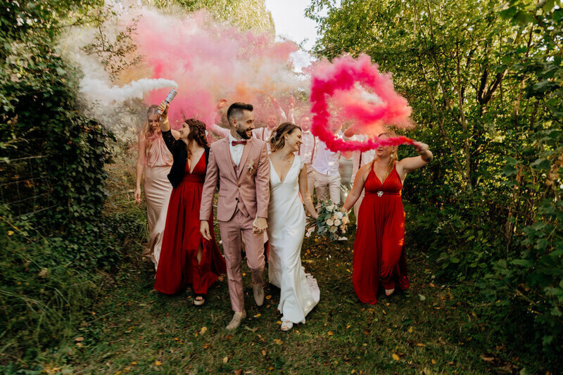 Mariés entourés de leurs demoiselles d'honneur dans une robe rouge et agitant des fumigènes roses en pleine nature pour la séance photo mariage en vendée.