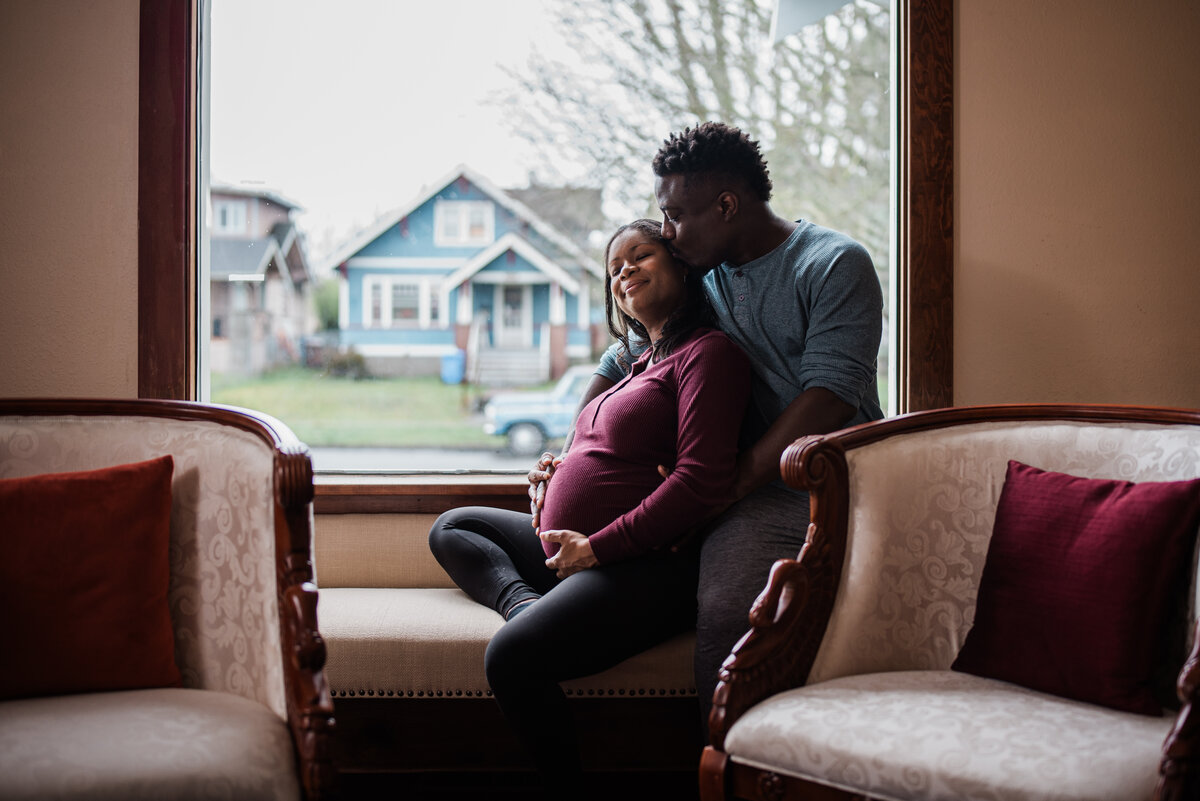Couple snuggling in the window seat of their front room, taking pregnancy photos at home for a cozy and relaxed vibe
