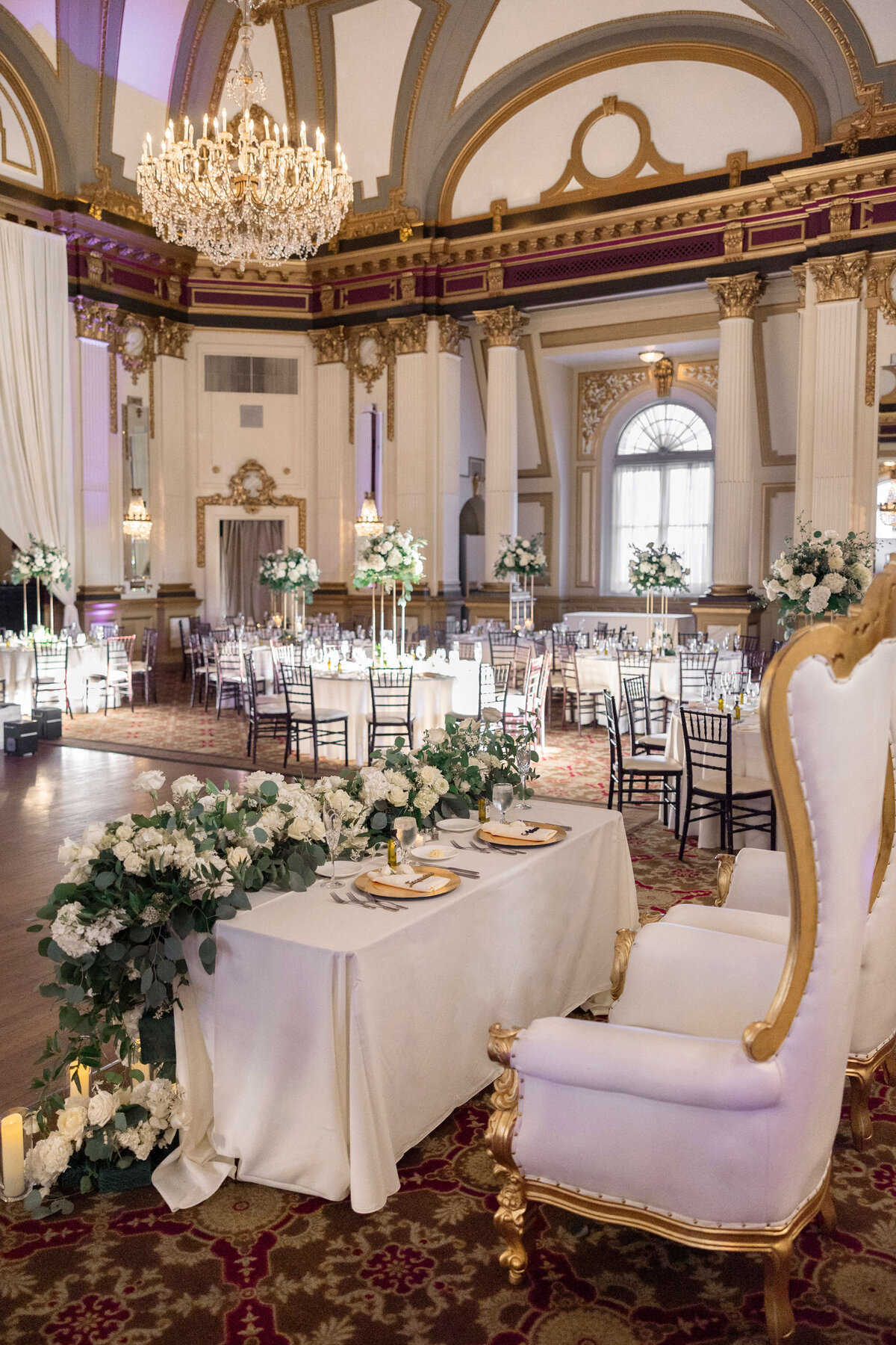 A lavish and elegant ballroom set for a wedding reception, featuring a long table adorned with white flowers and greenery. A white and gold ornate chair is prominent in the foreground. Chandeliers and tall floral arrangements enhance the grandeur.