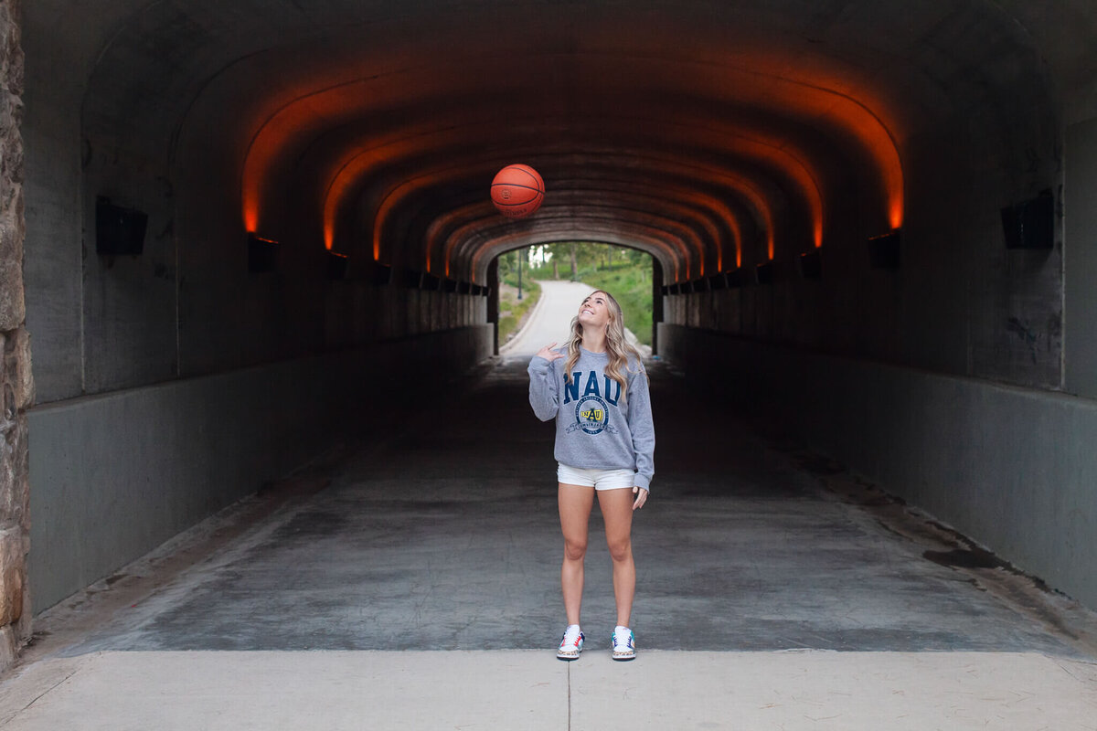 A person stands in a tunnel, balancing a basketball on their finger. They are wearing a gray sweatshirt and white shorts. The tunnel is lit with orange lights, and a path stretches out in the background.