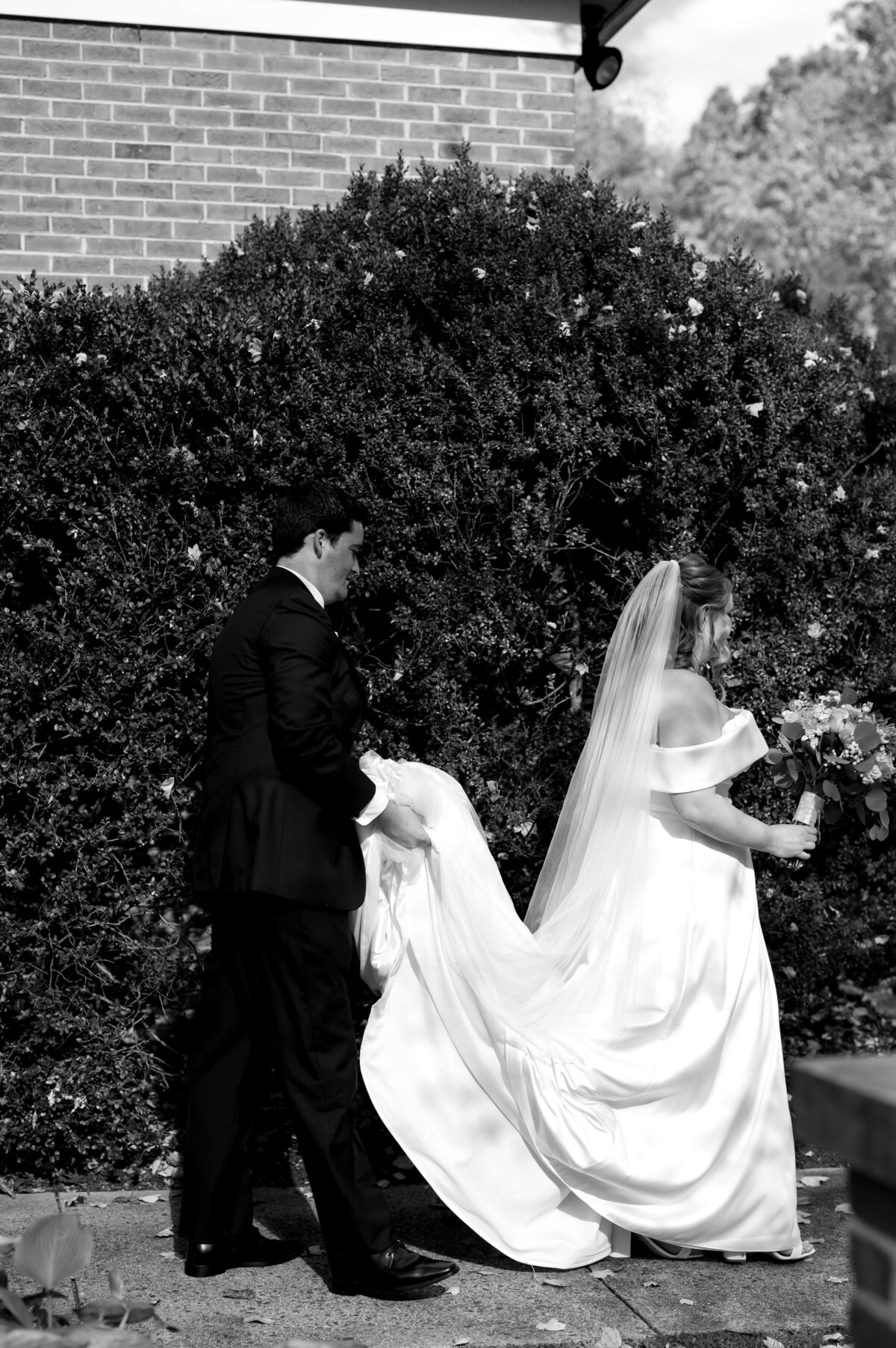 black and white candid wedding picture with groom carrying brides wedding dress as they walk through a garden together at middleburg community center for their Richmond wedding