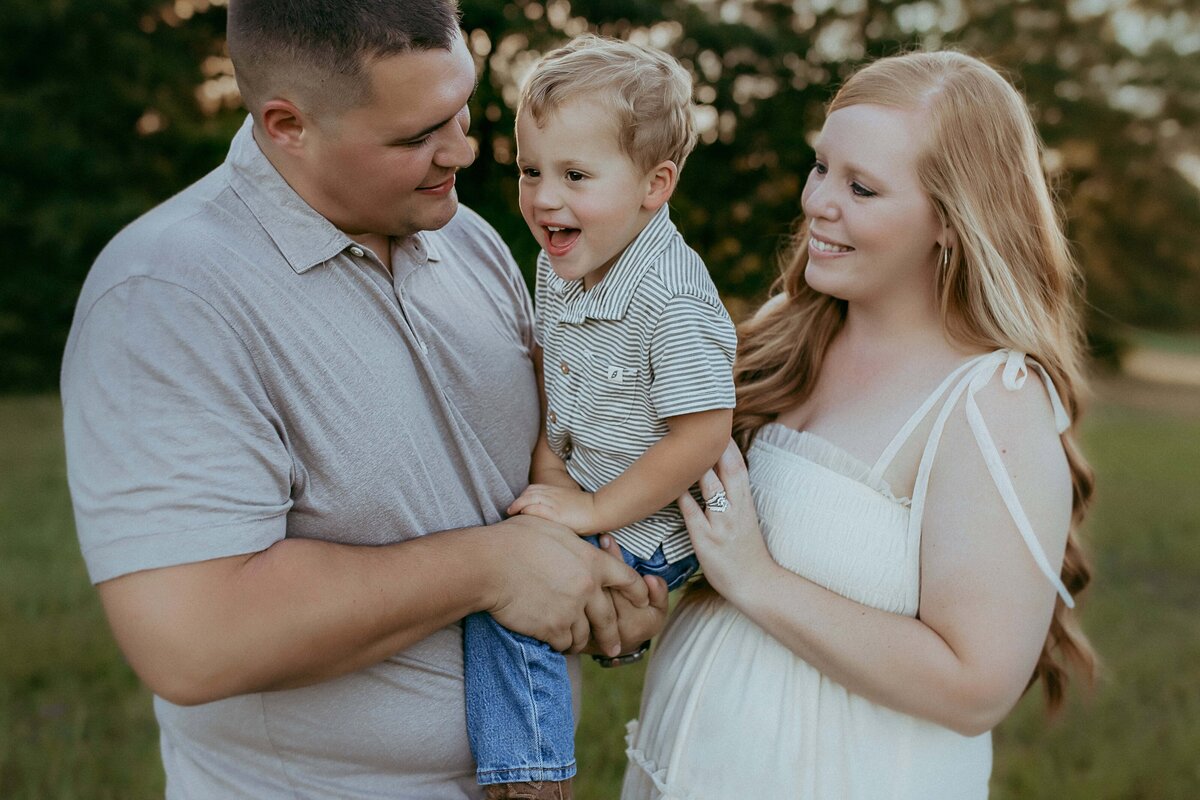 A man and woman stand outdoors holding a smiling young child, capturing a perfect family moment. The man wears a gray shirt, the woman a white dress, and the child a striped shirt. Surrounded by trees in the background, their joy is beautifully captured by a talented Warner Robins photographer.