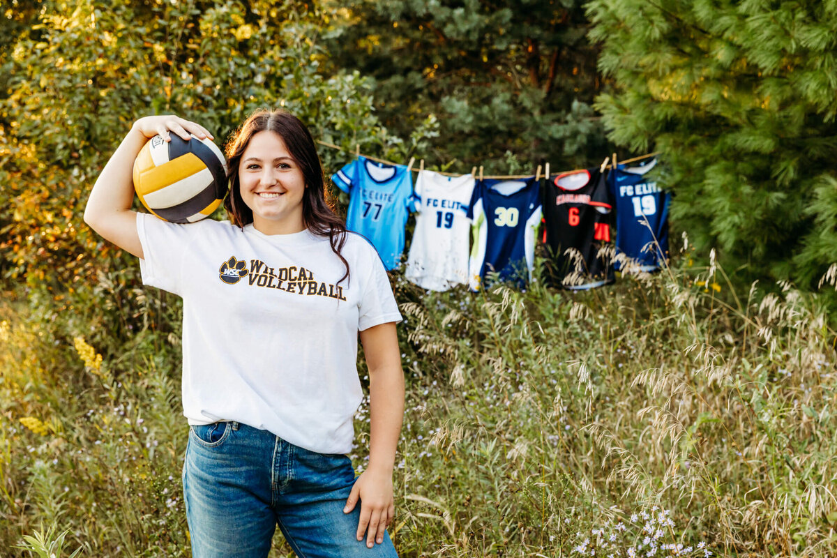 pretty high school girl in a white tshirt holding her volleyball on her shoulder as she stands in front of her other jerseys captured by Ashley Kalbus Photography