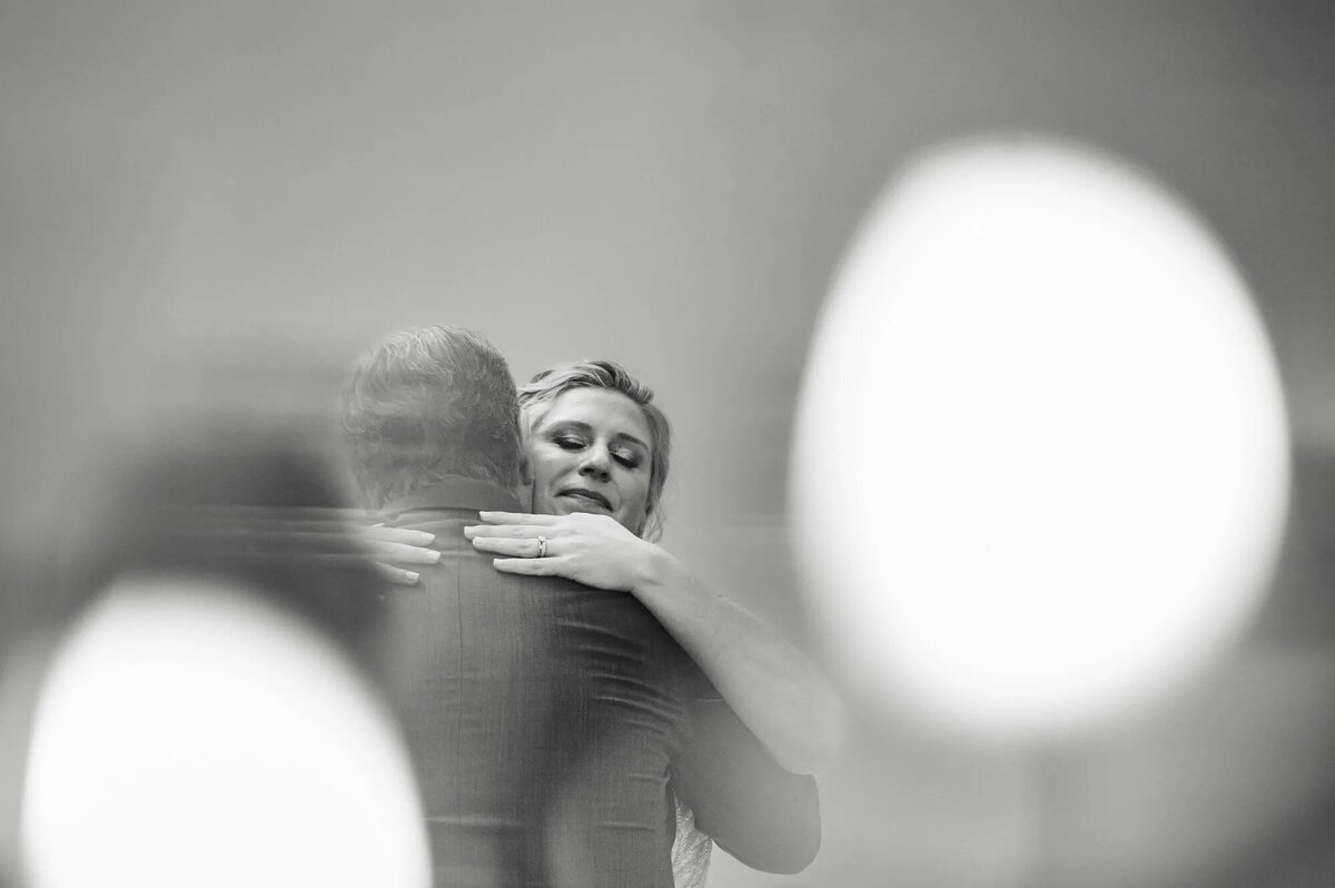 An intimate photo of a couple embracing, taken over the shoulder of one partner, with the focus on the other’s peaceful expression, and softly blurred lights in the foreground.
